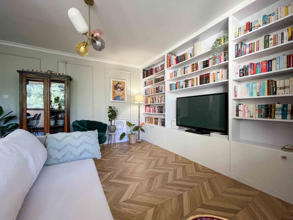 salon z dużą biblioteczką, jasnoszarą kanapą, drewnianym kredensem i dodatkami